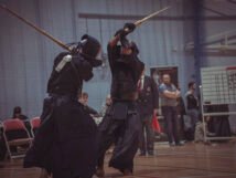 Two practitioners in traditional Kumdo attire engage in a sparring match with bamboo swords in a sports hall, surrounded by an audience observing the competition.