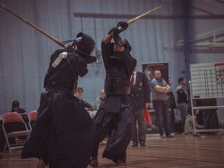 Two practitioners in traditional Kumdo attire engage in a sparring match with bamboo swords in a sports hall, surrounded by an audience observing the competition.